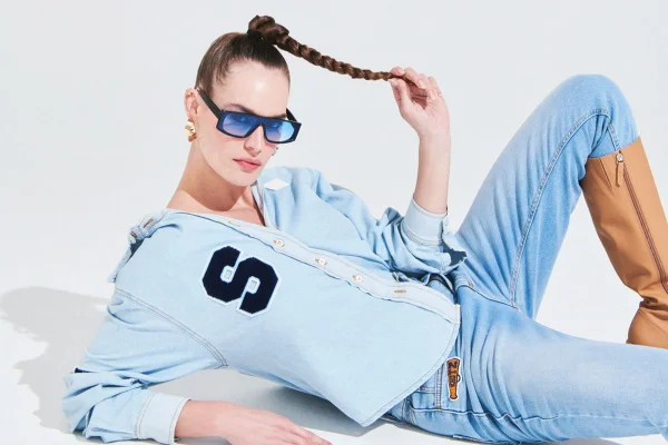 Modelo deitada segura uma trança no cabelo e usa look jeans com bota marrom