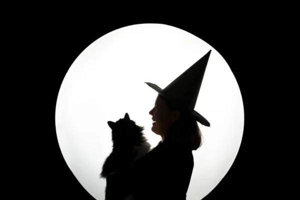 Imagem colorida de mulher fantasiada de bruxa carregando um gato