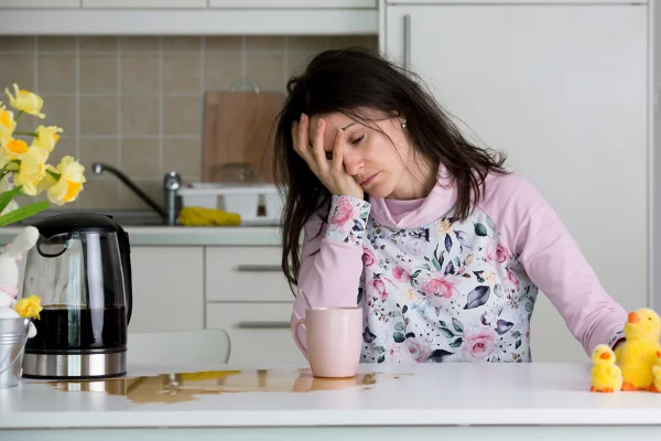 Foto colorida de mulher com a mão no rosto. Ela está de pijama e perto de uma bancada em que há café derrubado - Metrópoles