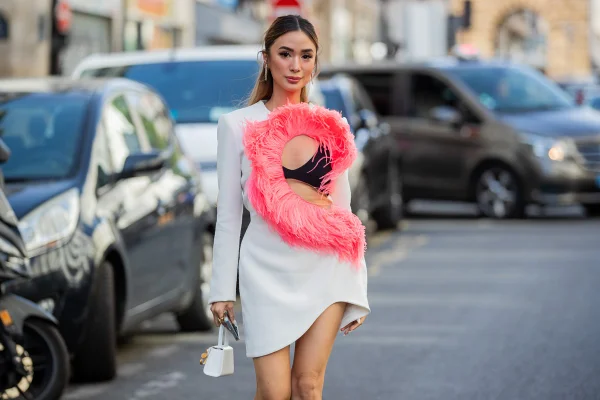 Mulher branca, jovem e asiática com cabelos lisos andando pelas ruas de Paris durante a Semana de Alta-Costura. Ela usa um sutiã preto e um vestido curto branco com um recorte no busto. Esse decote tem plumas rosas costuradas.