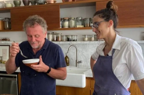 A apresentadora e cozinheira argentina Paolla Carosella ao lado de seu ex-marido Jason Lowe em sua cozinha. O homem prova comida em uma tigela e ela olha, sorrindo - Metrópoles