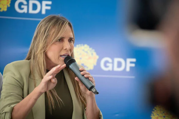 Foto colorida de mulher de cabelos loiras e de roupas falando com microfone na frente de fundo com logo do Governo do Distrito Federal