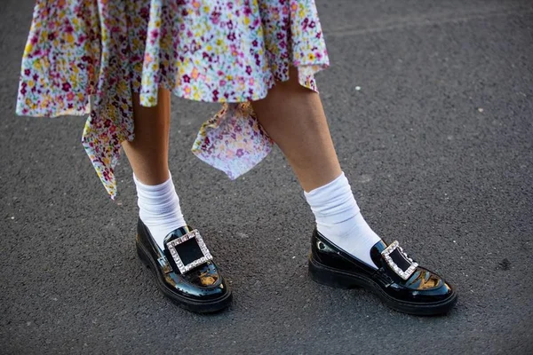 Pessoa andando na rua com um vestido esvoaçante florido, sapato mocassim preto e meia branca. Na foto é possível ver apenas as pernas e os pés da pessoa.