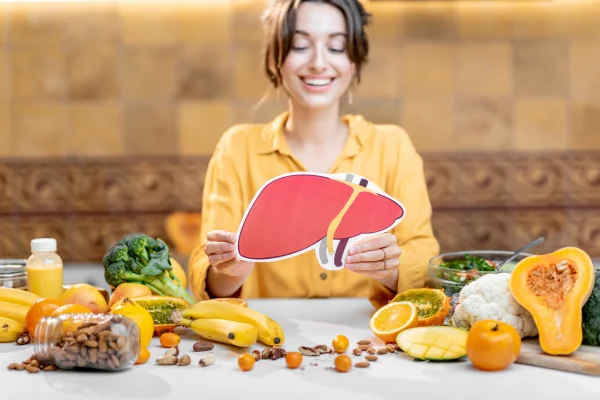 Foto colorida de mulher com blusa amarela, segurando um desenho que imida um órgão humano, rodeada de alimentos sobe uma mesa. - Metrópoles.