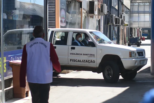 Imagem colorida de um funcionário da Diretoria de Vigilância Sanitária, da Secretaria de Saúde do Distrito Federal. O homem veste um colete branco e um casaco vermelho. Ao fundo, uma viatura de cor branca, do mesmo órgão