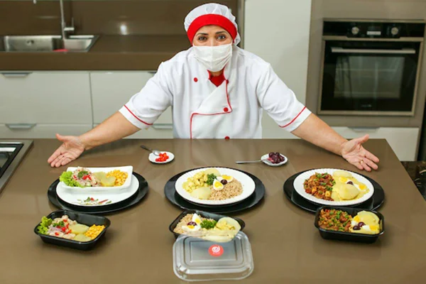 Cozinheira de dolmã, touca e máscara atrás de balcão com pratos peruanos