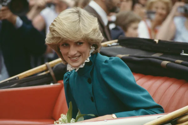 Princesa Diana era branca, cabelo loiros curtos e olhos claros. Ela está sentada no carro oficial da realeza com um vestido verde escuro.