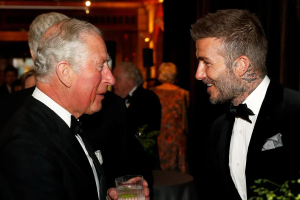 Foto colorida - Dois homens brancos se olhando e sorrindo. Na direita, Rei Charles, com uma aparência mais velha e, na esquerda, David Beckham, mais jovem e com tatuagens no pescoço