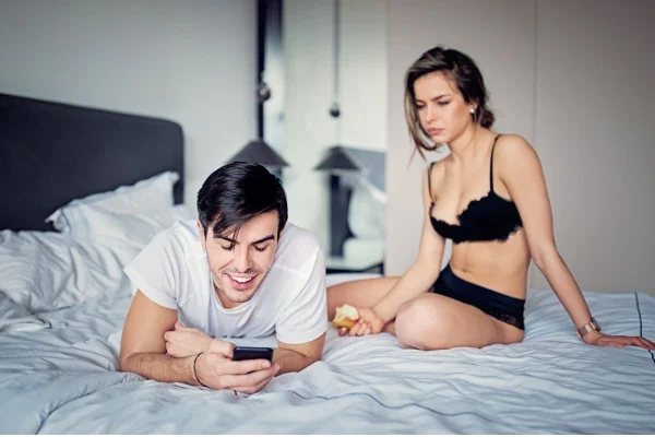 casal heterossexual branco em uma cama e o homem está olhando o celular