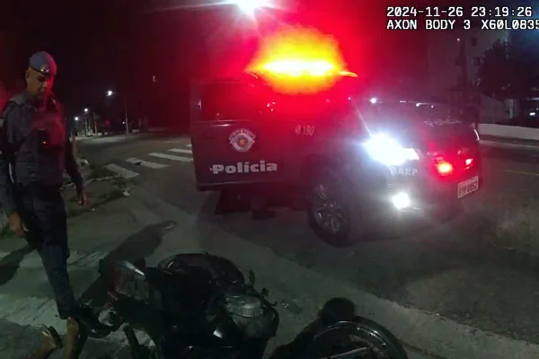 Imagem colorida mostra policial chutando arma para debaixo de morto. ao lado uma viatura está com a sirene ligada. É noite - Metrópoles