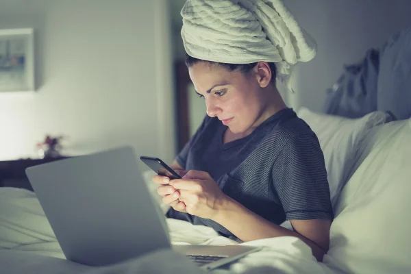 Jovem mulher com toalha na cabeça deitada em cama com o celular na mão e o laptop aberto - Metrópoles
