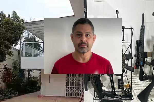 Foto centralizada de homem branco, cavanhaque grisalho e camiseta vermelha. Ao fundo, esquerda, casa de dois andares branca. Direita, fuzis apoiados em parede branca - Metrópoles