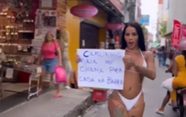 Mulher de biquíni branco desfila na rua com cartaz na mão - Metrópoles