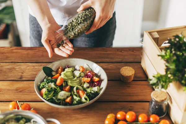 Foto colorida de mão segurando vidro e a outra tem dois grãos esverdeados. Em uma mesa, há salada e vegetais - Metrópoles