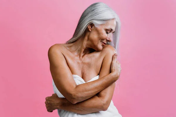Foto colorida de uma mulher branca com o cabelo grisalho enrolada em uma toalha branca e se abraçando em um fundo rosa - Metrópoles