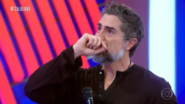 Na imagem colorida, um homem está posicionado no centro. Ele tem cabelos curtos e grisalhos e usa camiseta preta