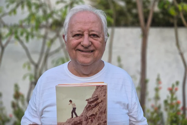 Imagem colorida de Caio Tibúrcio sorrindo, segurando livro e de blusa branca - Metrópoles
