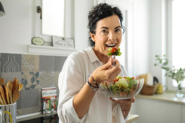 Foto colorida de mulher comendo salada. Ela está em uma cozinha - Metrópoles