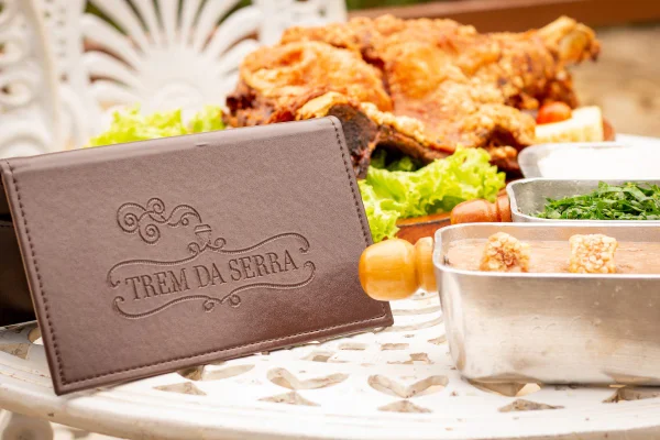 Restaurante Trem da Serra. Na foto, uma mesa com um cardápio escrito Trem da Serra, travessas de tutu de feijão, couve, arroz e de leitão a pururuca - Metrópoles
