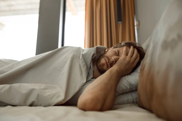 Homem deitado na cama fechando os olhos e colocando a palma da mão no rosto demonstrando estar cansado. Metrópoles