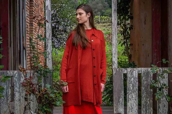 Mulher branca e jovem, de cabelo liso castanho, posa para foto em uma casa de madeira na serra gaúcha. Ela usa um look todo vermelho com calça, blusa, casaco e cardigan por cima. - Metrópoles