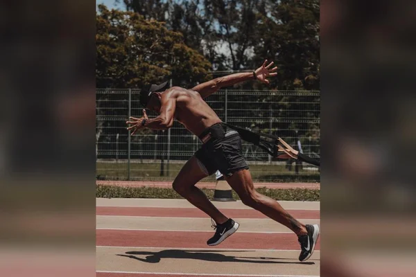Na imagem colorida, um homem está posicionado no centro. Ele tem cabelos curtos e escuros. Ele está sem blusa, usa roupa de atleta e está em posição para correr