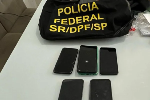 Imagem colorida mostra cinco celulares sobre superfície branca e bolsa preta de tecido com identificação da Polícia Federal - Metrópoles