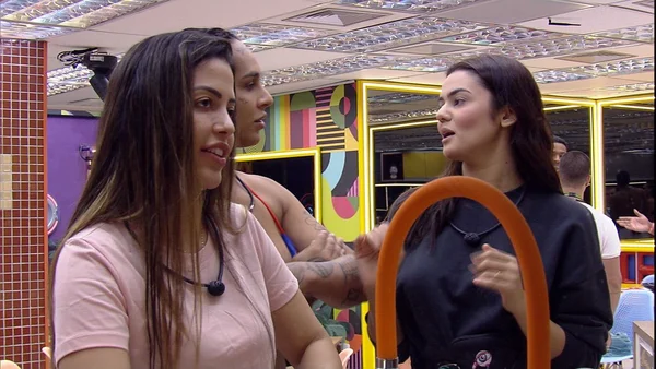 Três mulheres conversam em meio à cozinha. Em primeiro plano, uma com blusa rosa, e, em segundo, uma de blusa preta.