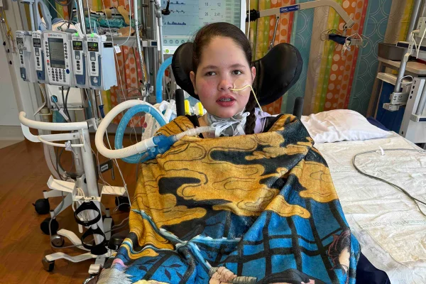 Foto colorida de adolescente em ambiente hospitalar, conectada a tubos, sentada em cadeira de rodas - Metrópoles.