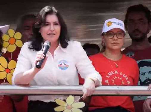 Simone Tebet discursa ao lado da mulher de Lula, Janja, em campanha em Minas Gerais. Ambas estão em cima de carro de som, cercadas por outras pessoas, e seguram flores de papel que simbolizam campanha de luta contra pedofilia - Metrópoles