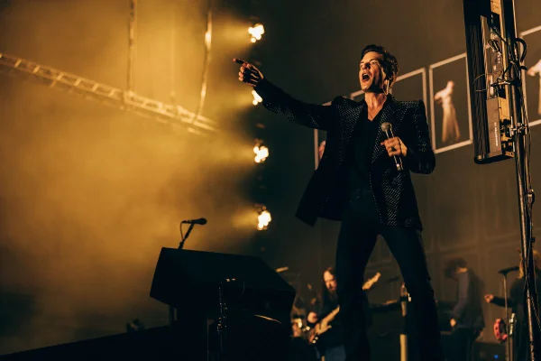 Brandon Flowers, vocalista da banda The Killers, canta no palco segurando um microfone e apontando para a plateia. Ao fundo, uma luz amarela - Metrópoles