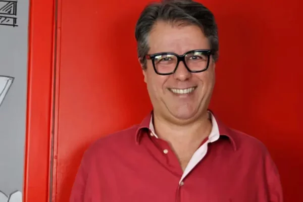 Tony Goes é clicado sorridente, usando óculos e uma camisa vermelha - Metrópoles