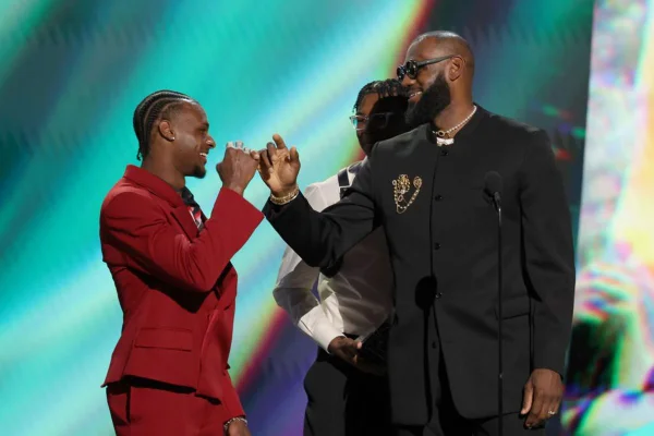 Imagem colorida mostra Bronny James, um adolescente negro, com terno vermelho, cumprimentando o pai, LeBron James, que está de terno preto, em um palco - Metrópoles