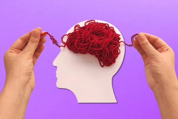 Foto colorida com fundo rosa de desenho em cor branca que imita a face humana e uma bola de lã de cor vermelha que simula a existência de um cérebro. - Metrópoles.