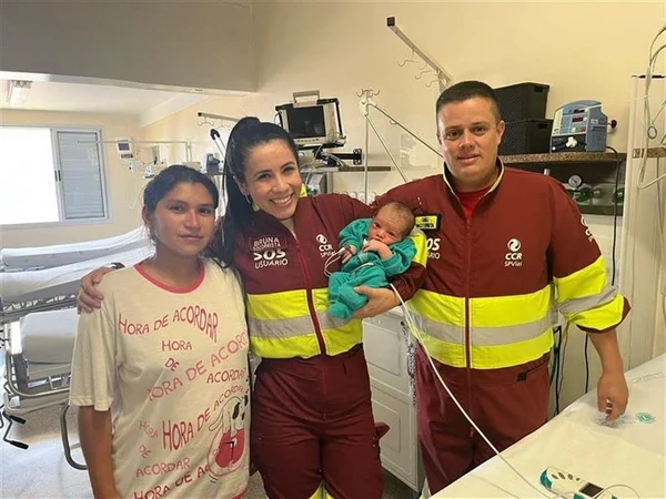 Imagem colorida mostra um bebê no colo de uma socorrista, mulher branca vestida com um uniforme vermelho, ao lado de outro socorrista, um homem branco com o mesmo uniforme, e a mãe da criança, uma mulher de pele parda vestindo uma camiseta branca - Metrópoles