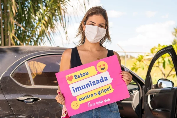 Mulher com máscara branca segurando placa rosa em frente a carro preto. Céu azul e folha de palmeira ao fundo