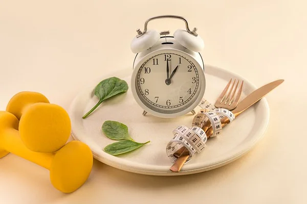 prato post sob uma mesa com halteres de musculação ao lado. Em cima do prato há um relógio, folhas de manjericão e talheres enrolados em uma fita métrica