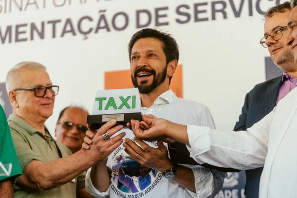 Imagem colorida mostra ao fundo Ricardo Nunes, homem branco, de cabelo e barba pretos, falando ao microfone, com um sinal luminoso de carro de táxi em primeiro plano - Metrópoles