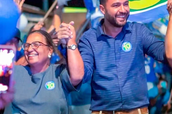 candidatos dão as mãos em eleição. Ambos com camisas azul. À esquerda, mulher mais velha. À direita, homem jovem