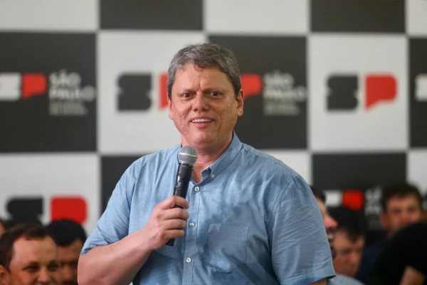 Imagem colorida mostra Tarcísio de Freitas, homem branco e grisalho, de camisa de mangas curtas azul, falando ao microfone com expressão sorridente, com um banner do governo de SP ao fundo - Metrópoles