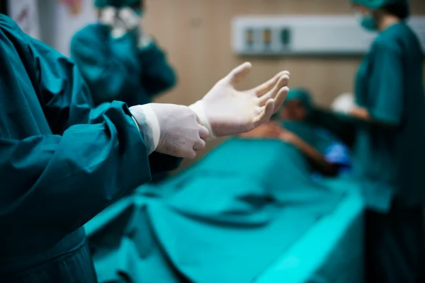foto colorida de centro cirúgico com médico em primeiro plano colocando luvas para dar início a cirúrgia - Metrópoles.