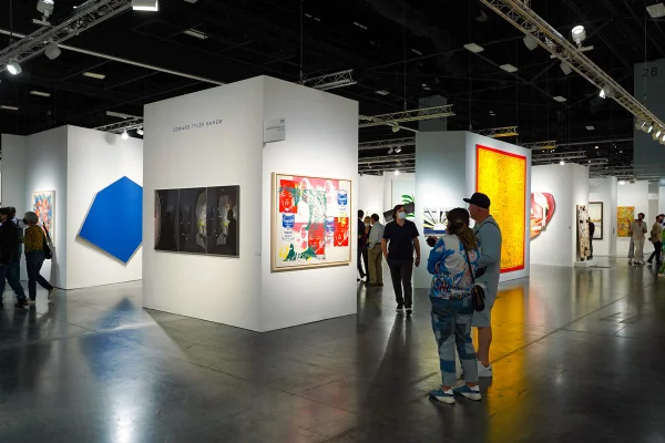 Um homem e uma mulher, ambos brancos e de meia idade, admiram uma obra em uma parede de uma exposição de arte. A foto é do evento Art Basel, em Miami, nos EUA, em 2021. Ela usa uma jaqueta e uma calça, ambos jeans, um boné e tênis. O homem usa um conjunto de shorts e casaco de moletom e tênis branco. - Metrópoles