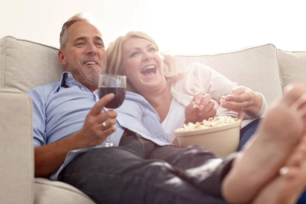 Casal sorri enquanto assiste algo, bebe vinho e come pipoca