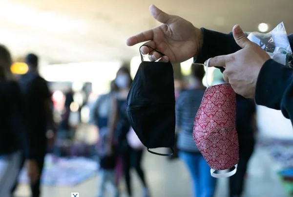 Mão segurando máscaras e pessoas passando ao fundo