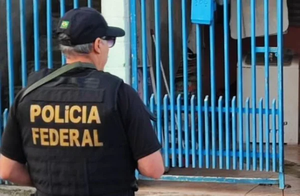 Homem de costas com colete da Polícia Federal e boné preto em frente a um portão azul