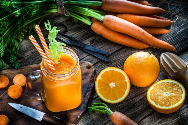 Bebida saudável: vista de alto ângulo de um copo cheio de suco orgânico fresco de laranja e cenoura, tirado em uma mesa de cozinha rústica de madeira. Laranjas e cenouras inteiras e fatiadas estão ao redor do copo. Uma faca de cozinha e uma tesoura de cozinha completam a composição. Metrópoles