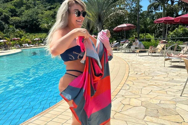 Simony posa de chinelo, biquíni preto e óculos escuros, segurando uma canga colorida - Metrópoles