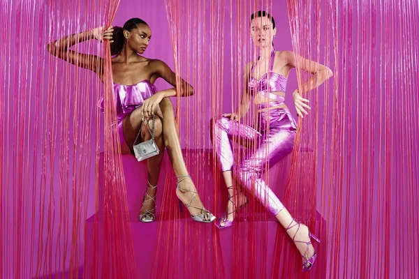 Duas modelos jovens, uma negra e uma branca, posando para foto na campanha de divulgação da nova coleção da Corello. Ambas usam roupas rosas metalizadas e acessórios também metalizados.
