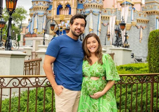 Homem com camiseta azul e bermuda bege ao lado de mulher com vestido verde, grávida, com a mão sobre a barriga - Metrópoles