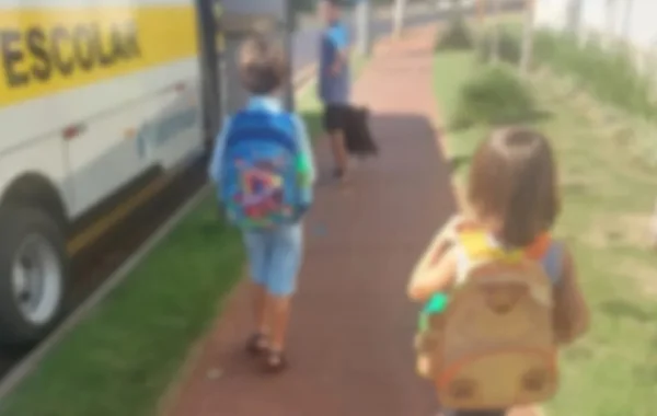 Duas crianças de costas com mochilas caminham em direção a uma van escolar